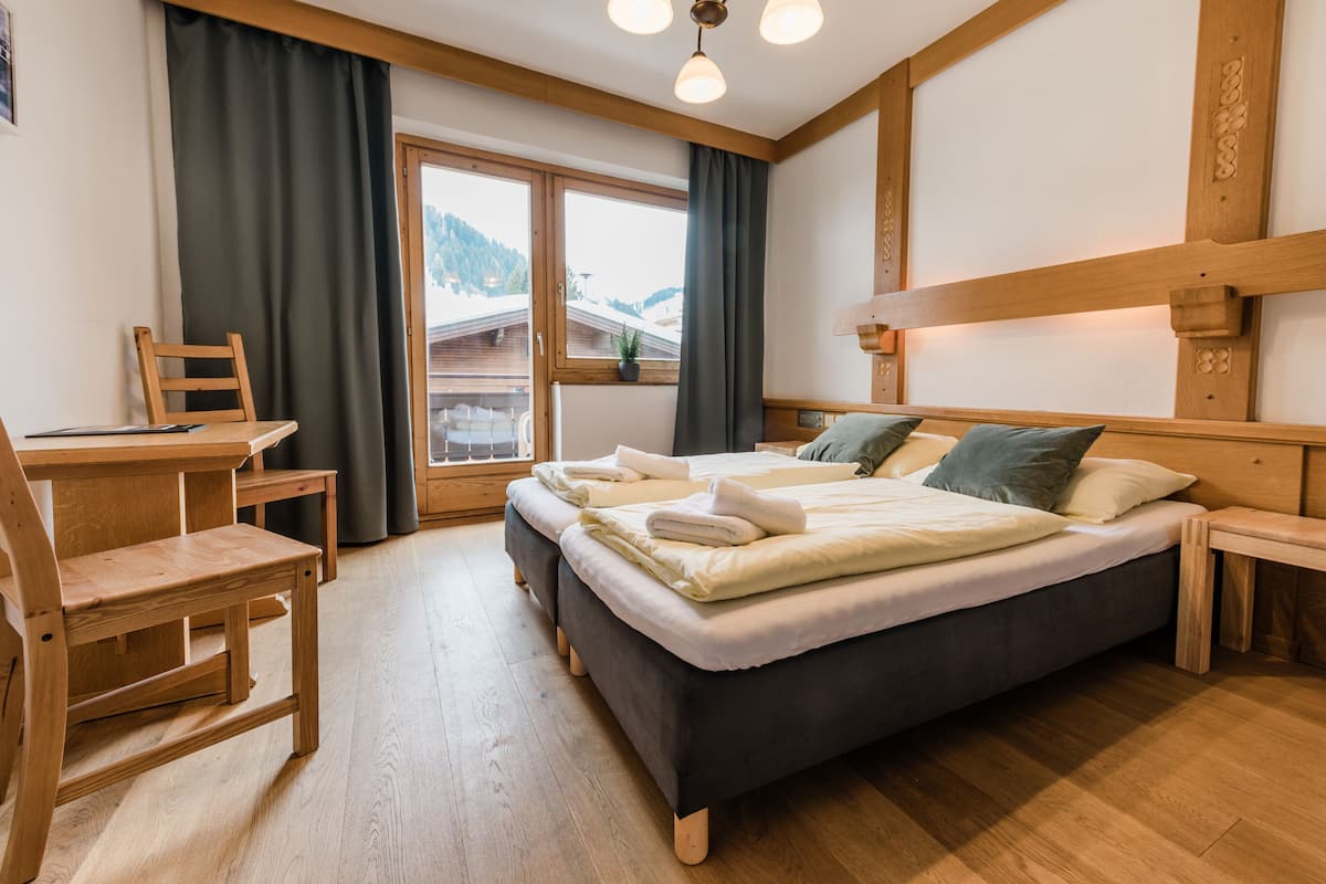 Een hotelkamer met een bed, een bureau en een stoel. Er is een raam met gordijnen, uitzicht op de bergen en een houten plafond.
