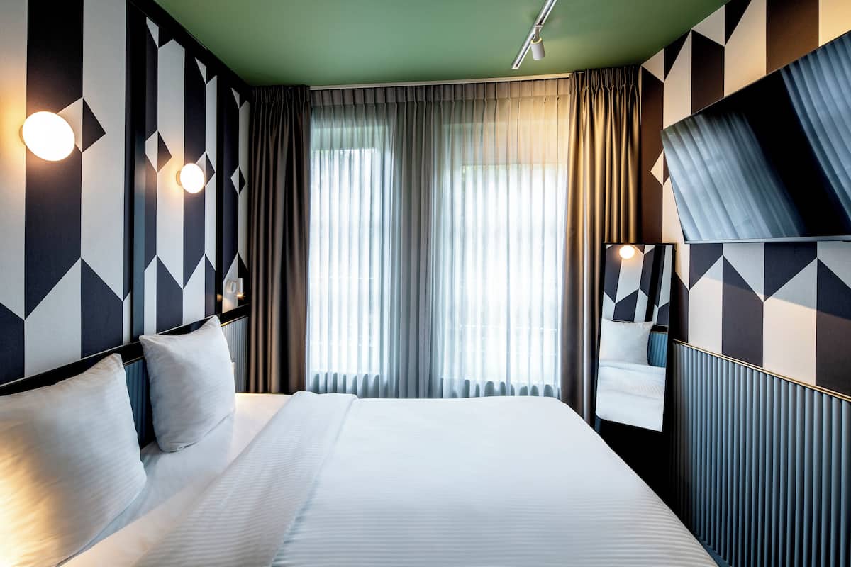 A hotel room with a large bed, a mirror, geometric-patterned walls, and a view of the outdoors through sheer curtains.