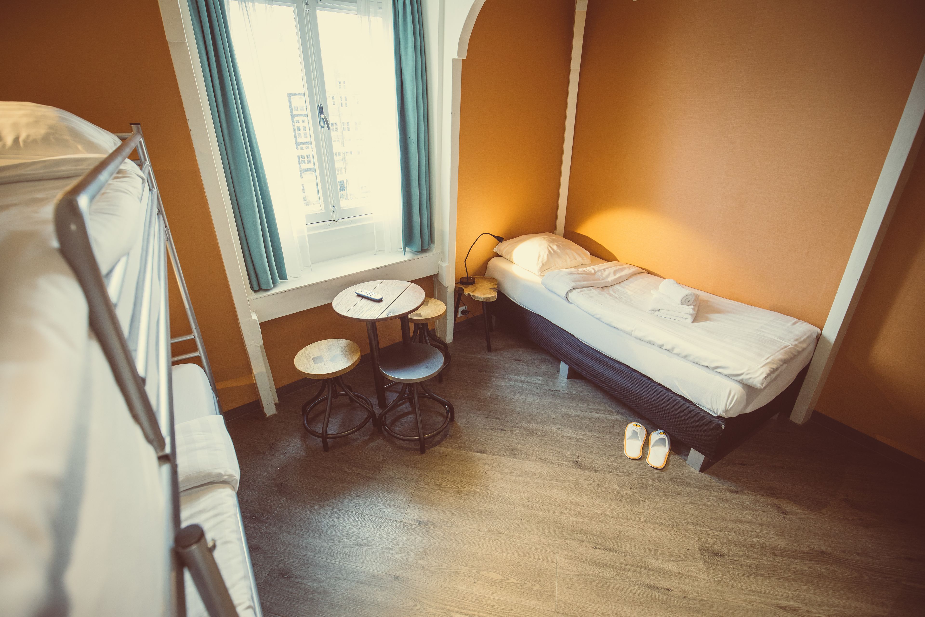 Ein kompaktes Hotelzimmer mit Etagenbett, einem Einzelbett, einem kleinen runden Tisch und einem Fenster mit Vorhängen.