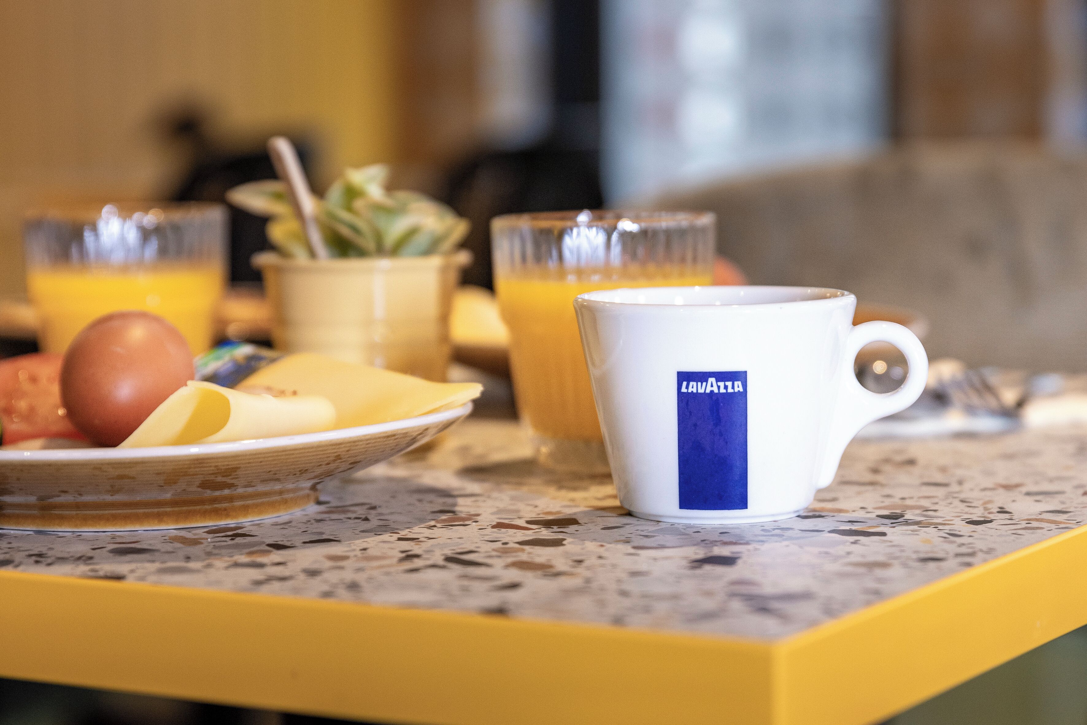 A breakfast setting with a plate of eggs, a bowl of fruit, and a mug labeled "Lavazza" on a speckled countertop.