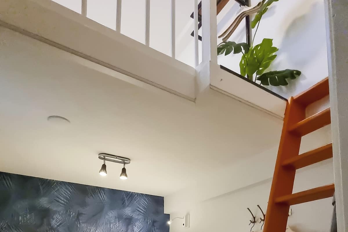 A dormitory room with two bunk beds, a wooden ladder, a small stool, a wall-mounted shelf, and a ceiling light fixture.