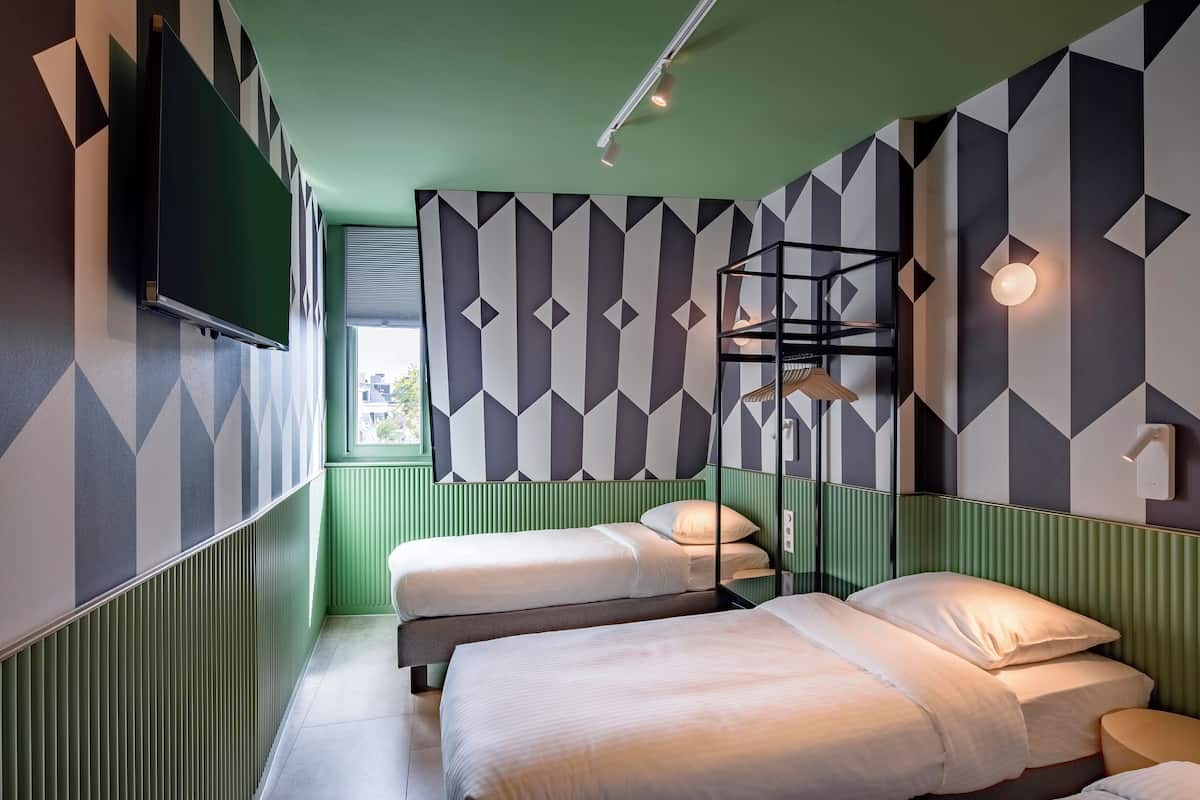 A room with two beds, a TV mounted on the wall, a shelf with a hanging lamp, and geometric patterned walls.