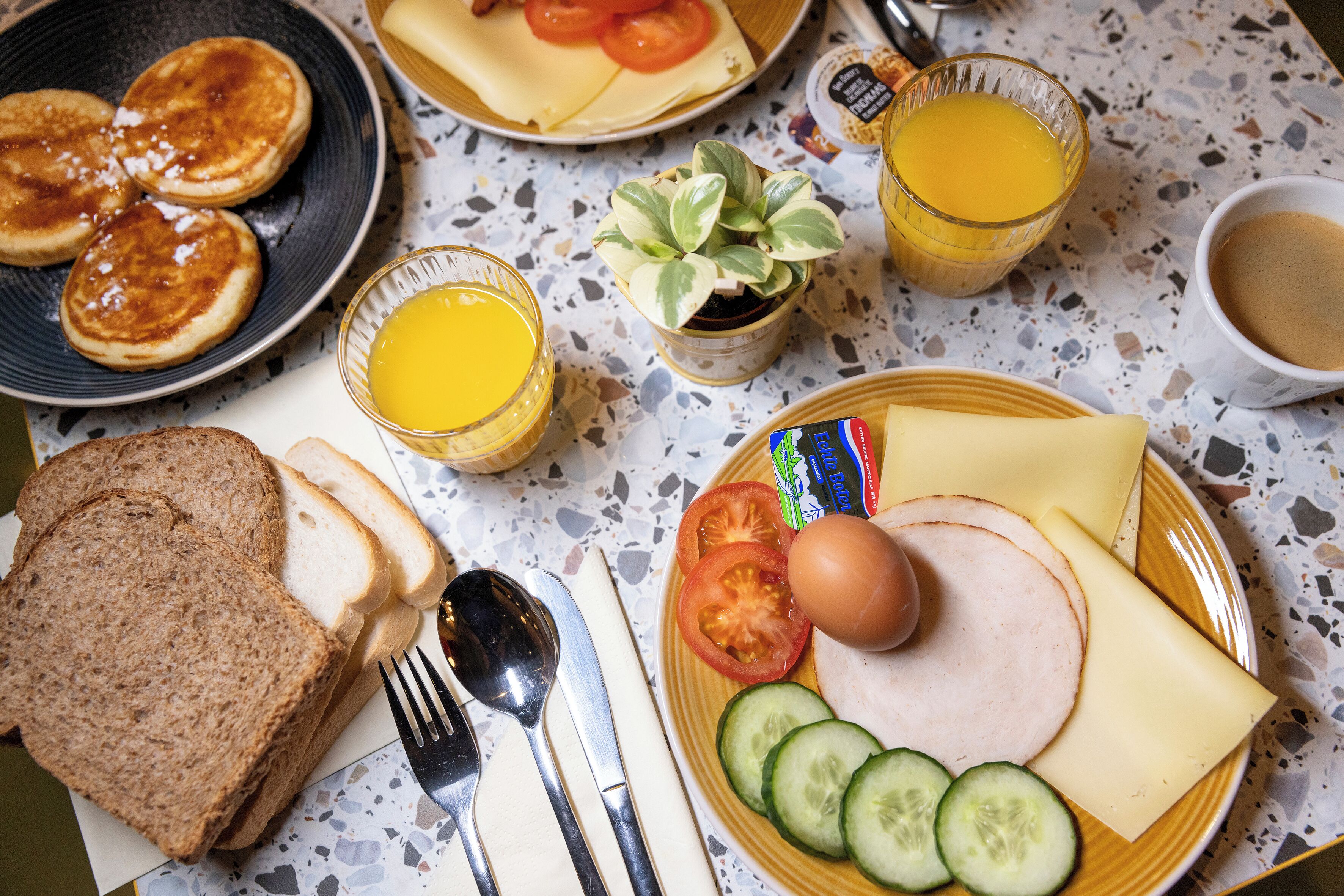 Une table de petit-déjeuner avec des crêpes, des toasts, des œufs, du fromage, des tomates, des concombres et du jus de fruits.