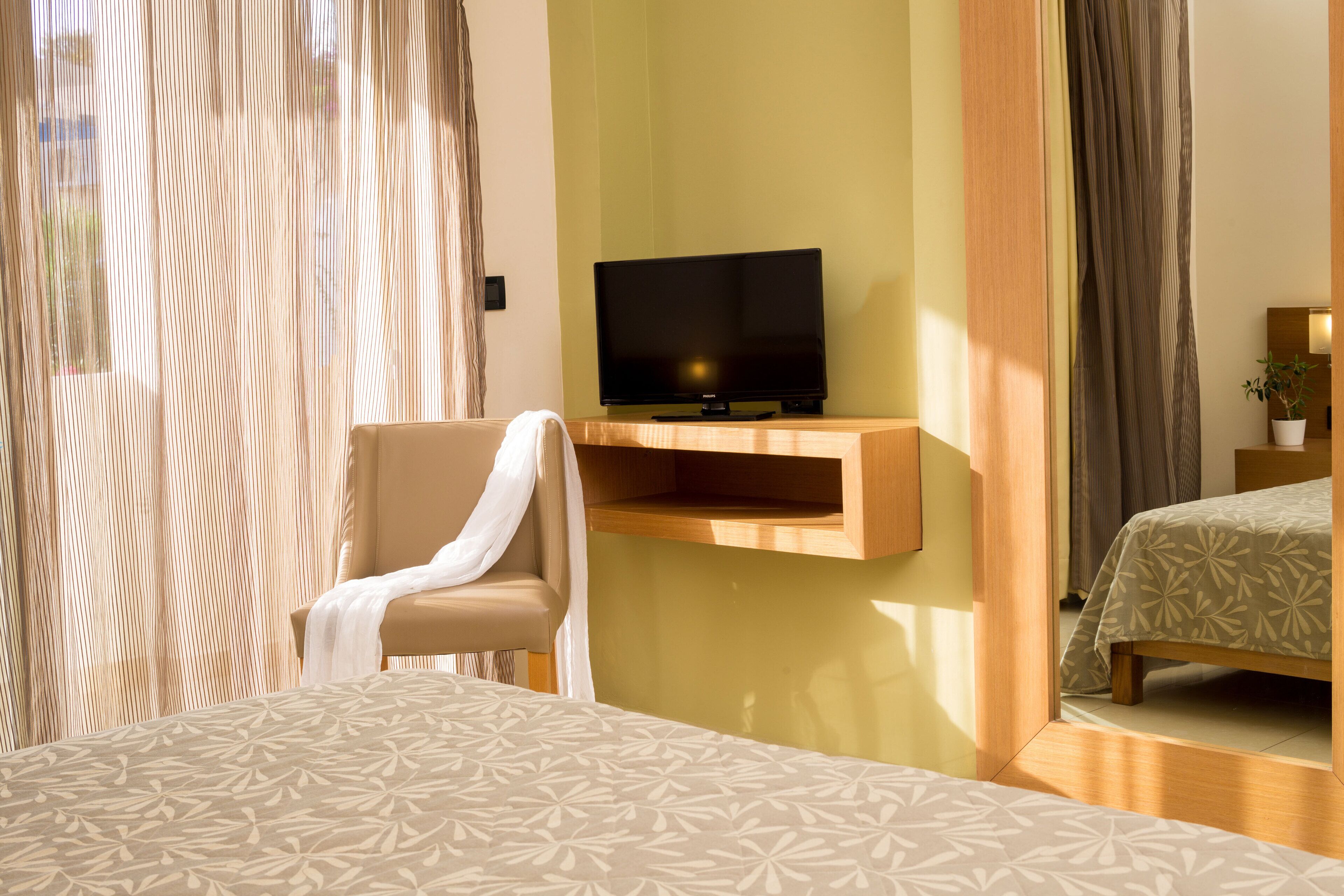 Una habitación de hotel con una cama, una silla, un televisor en un estante de madera y una planta en maceta.