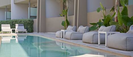 Zona de piscina con sillones reclinables y una mesa pequeña, rodeada de plantas grandes y un edificio con balcones.