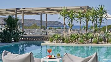 A poolside area with lounge chairs, a small table with a drink and fruit, and a view of the sea and mountains.