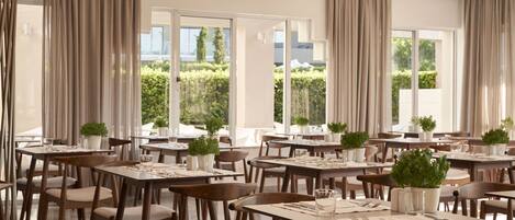 Un comedor amplio con varias mesas y sillas, ventanas grandes con cortinas y plantas en macetas.