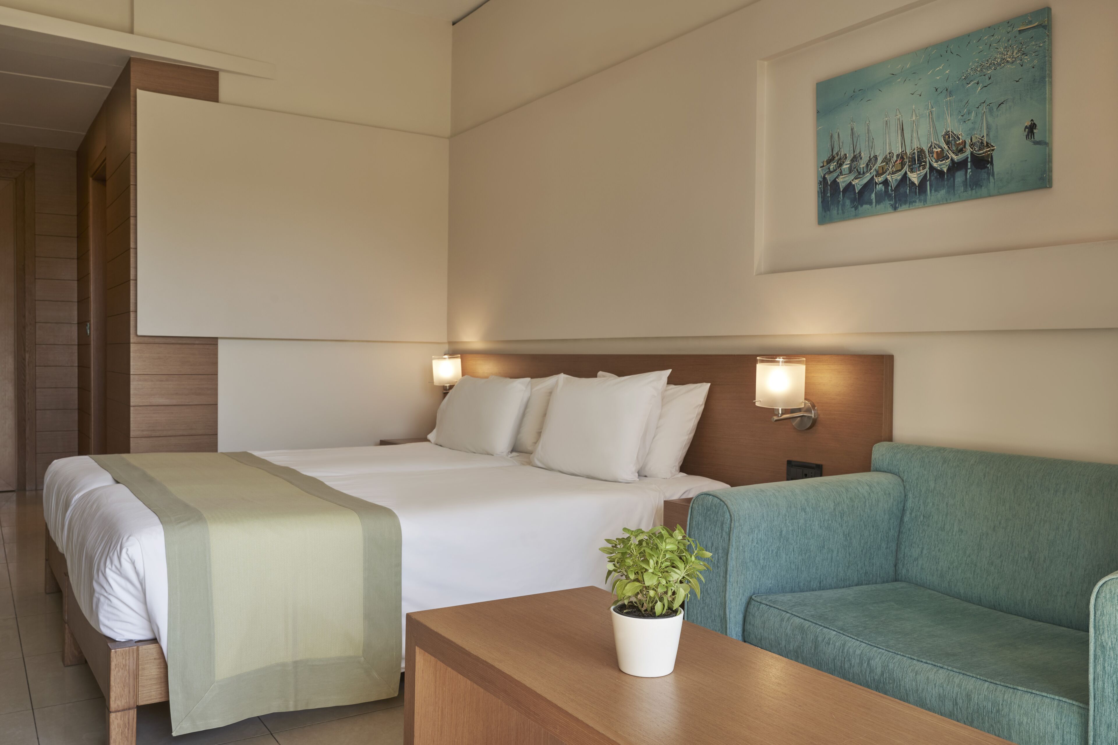 Une chambre d’hôtel avec un lit, un canapé, une table de chevet ornée d’une plante et un tableau accroché au mur.