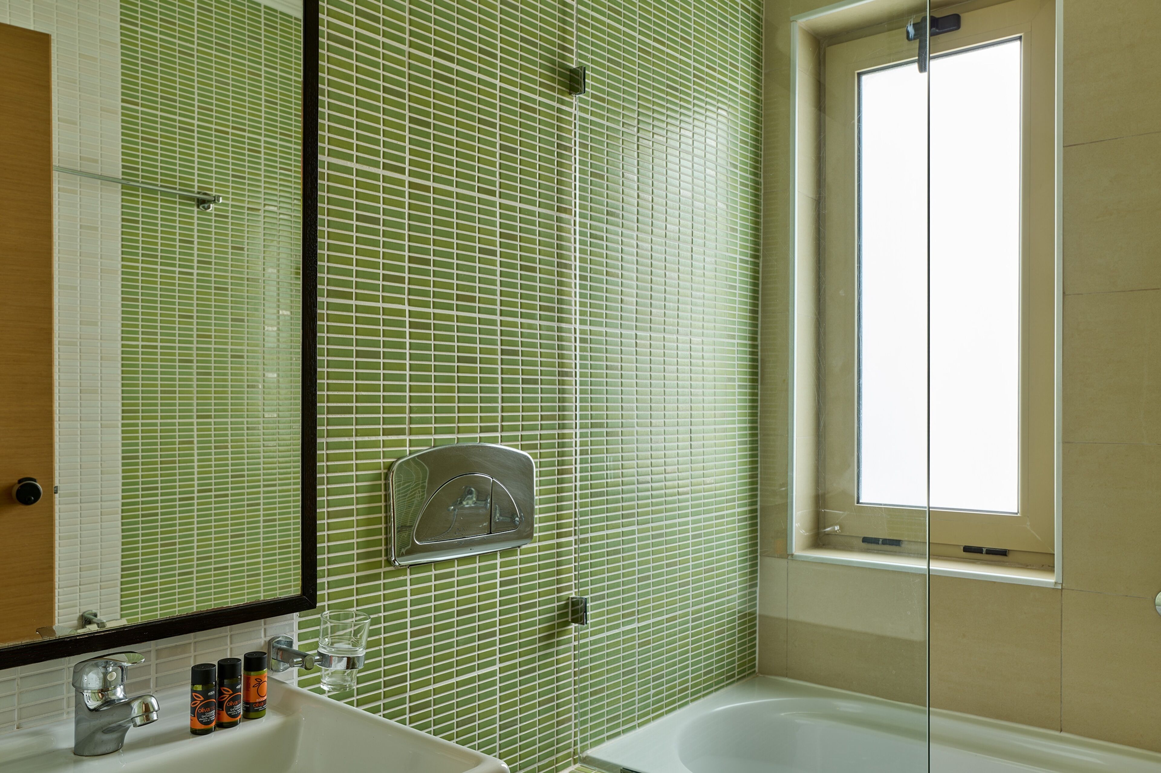 Un baño con paredes de azulejos verdes, un lavamanos blanco, un espejo y una ventana.