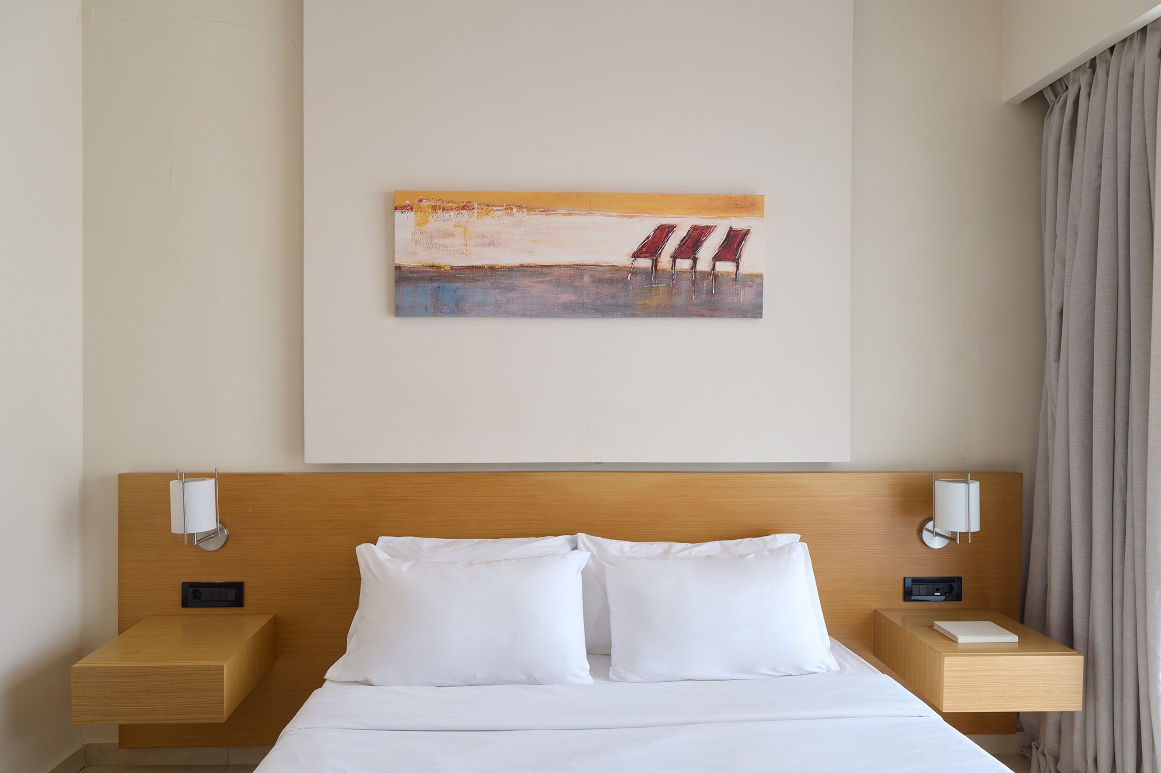 A hotel room with a wooden headboard, a bed with white linens, two bedside tables with lamps, and a painting of beach chairs on the wall.