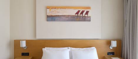 A hotel room with a wooden headboard, a bed with white linens, two bedside tables with lamps, and a painting of beach chairs on the wall.