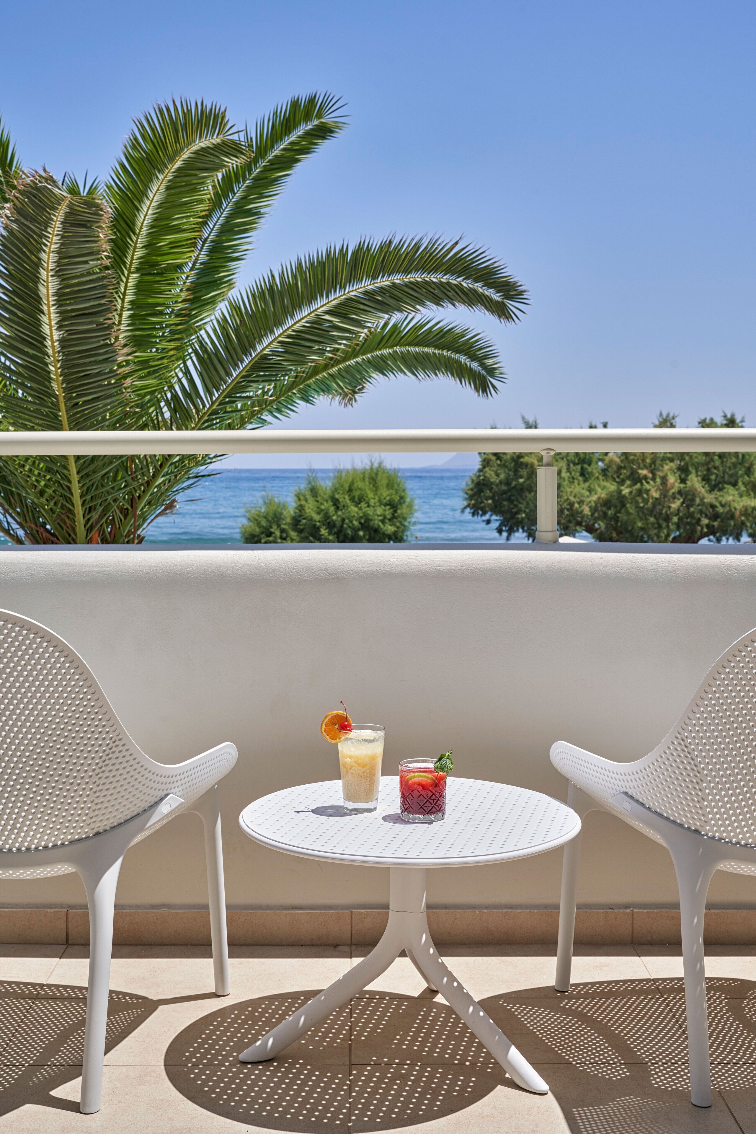 Un balcon avec deux chaises blanches et une petite table ronde, d’où l’on peut admirer une mer bleue et limpide ainsi que des palmiers.