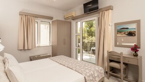 A bedroom with a bed, a desk, and a chair. There is a window with curtains, an air conditioning unit, and a television mounted on the wall. - Niriis Hotel (Chania)