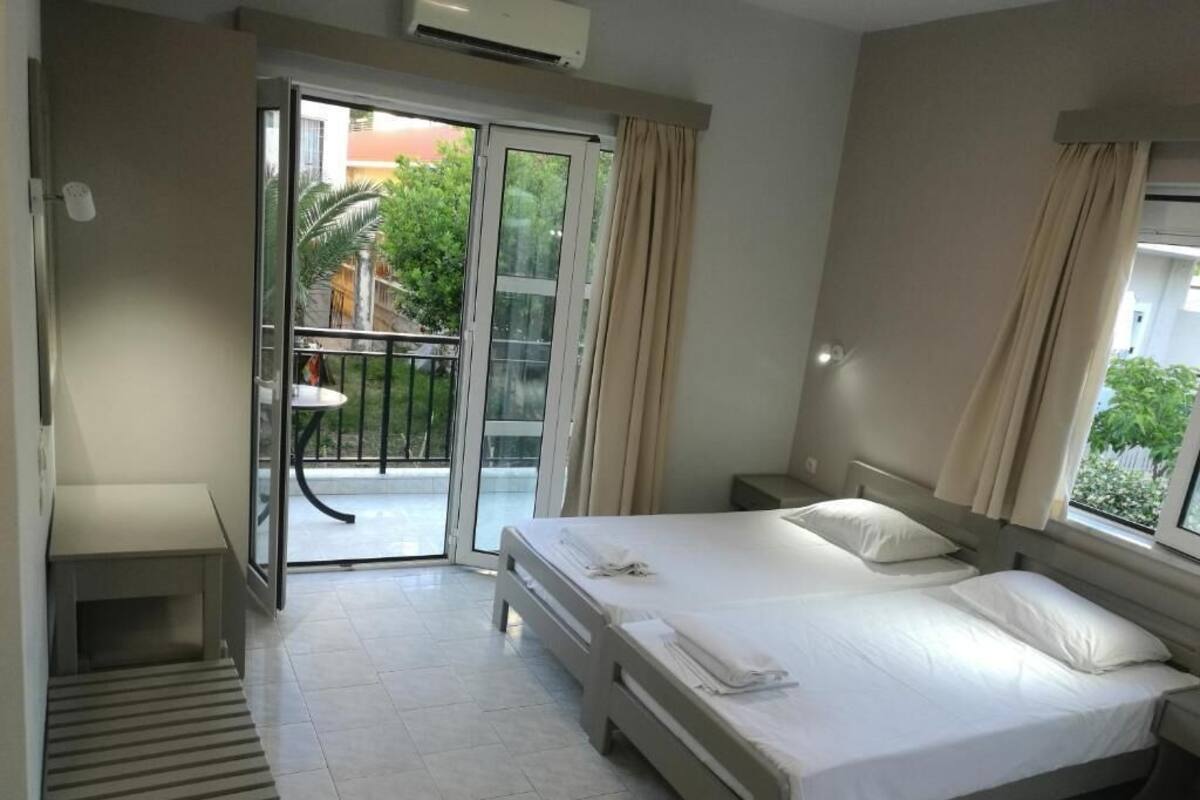 A hotel room with a bed, a small table, a bench, and a balcony with a view of greenery.