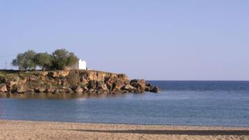 A coastal scene with a sandy beach, rocky cliffs, and a white building with trees.