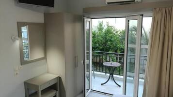 A hotel room with a bed, a desk, a mirror, a television, and a sliding glass door leading to a balcony with a table and chairs.