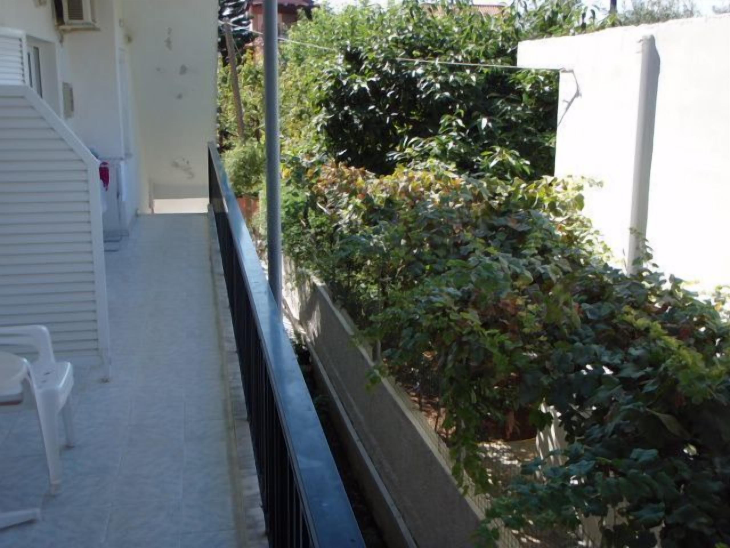 Un estrecho pasillo exterior con una pared blanca en un lado y una valla metálica en el otro. Se observa una silla de plástico blanca y un extintor rojo fijado a la pared.