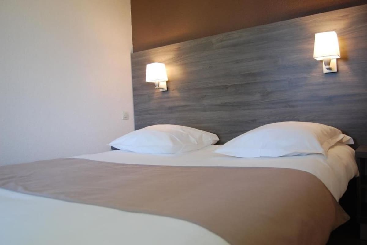 Habitación de hotel con cama, dos lámparas fijadas a la pared y cabecero de madera.