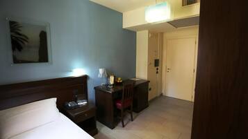 Ein Hotelzimmer mit einem Bett, einem Schreibtisch mit Stuhl, einer Lampe, einem kleinen Tisch mit Telefon und einer Vase sowie einer Tür zu einem weiteren Raum.