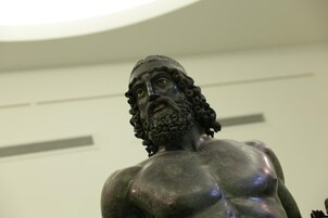 Una estatua de complexión musculosa y cabello rizado, exhibida en un interior con fondo neutro.