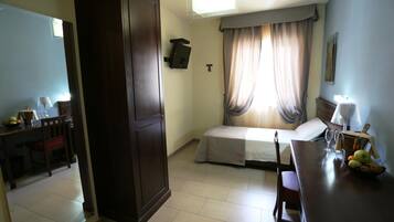 A hotel room with a bed, a desk with a chair, a wardrobe, a small table with fruit, and a window with curtains.