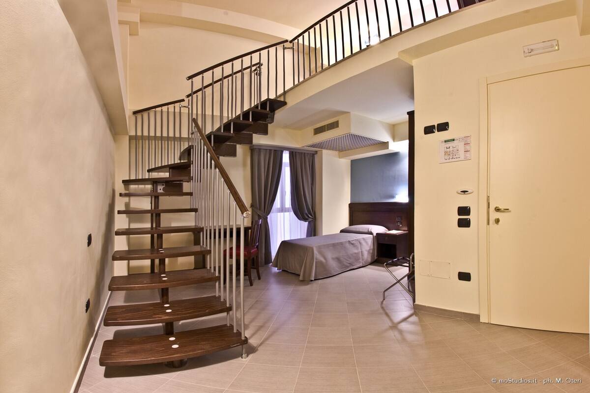 A hotel room with a staircase, a bed, a desk, and a door with a control panel.