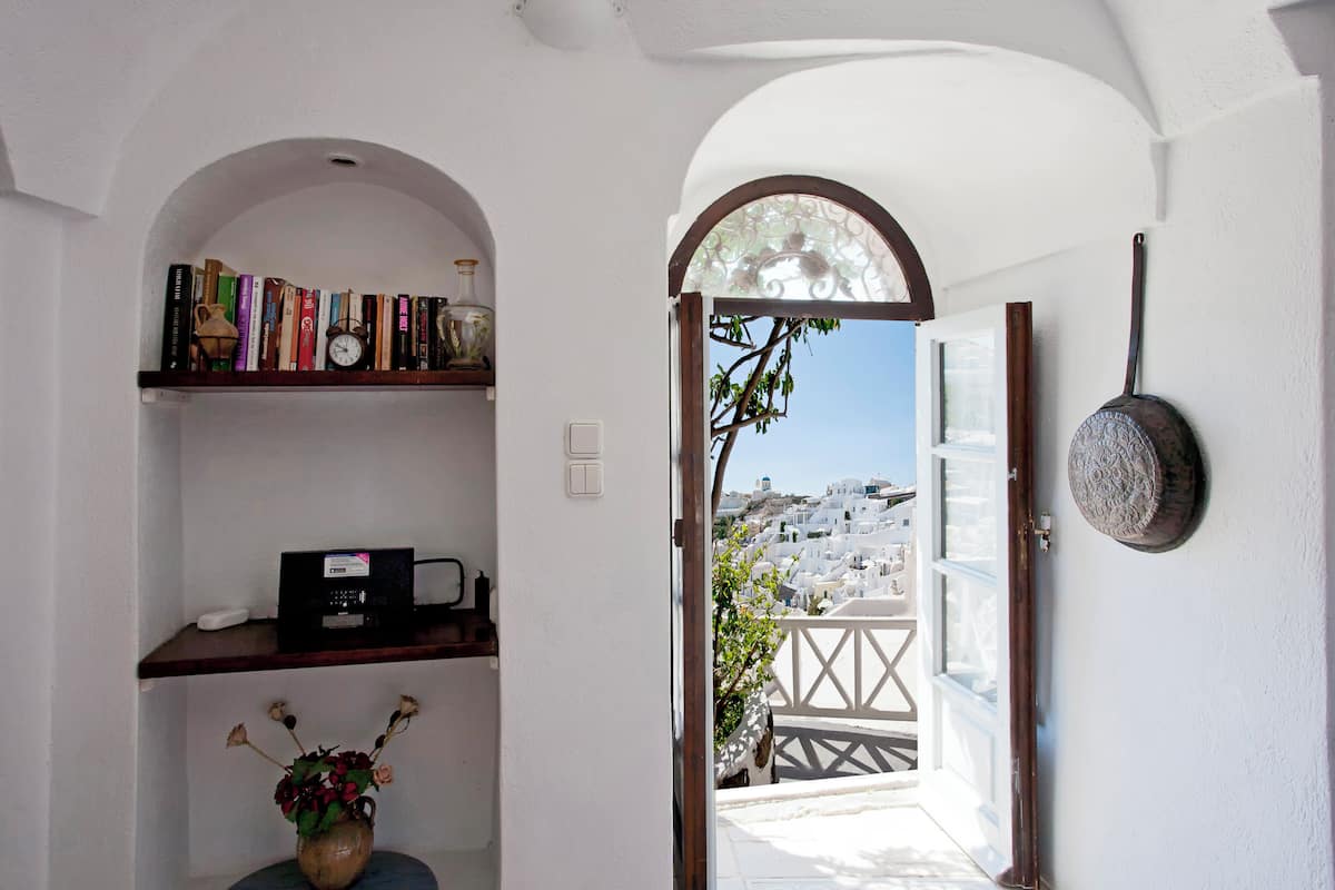 A doorway leading to a balcony with a view of white buildings, a shelf with books and a vase of flowers, and a wall-mounted pot.