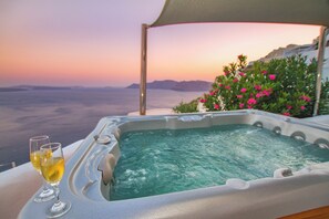 A hot tub with two glasses of wine, overlooking a scenic view with mountains and flowers. - Alexander's Suites (Santorini)
