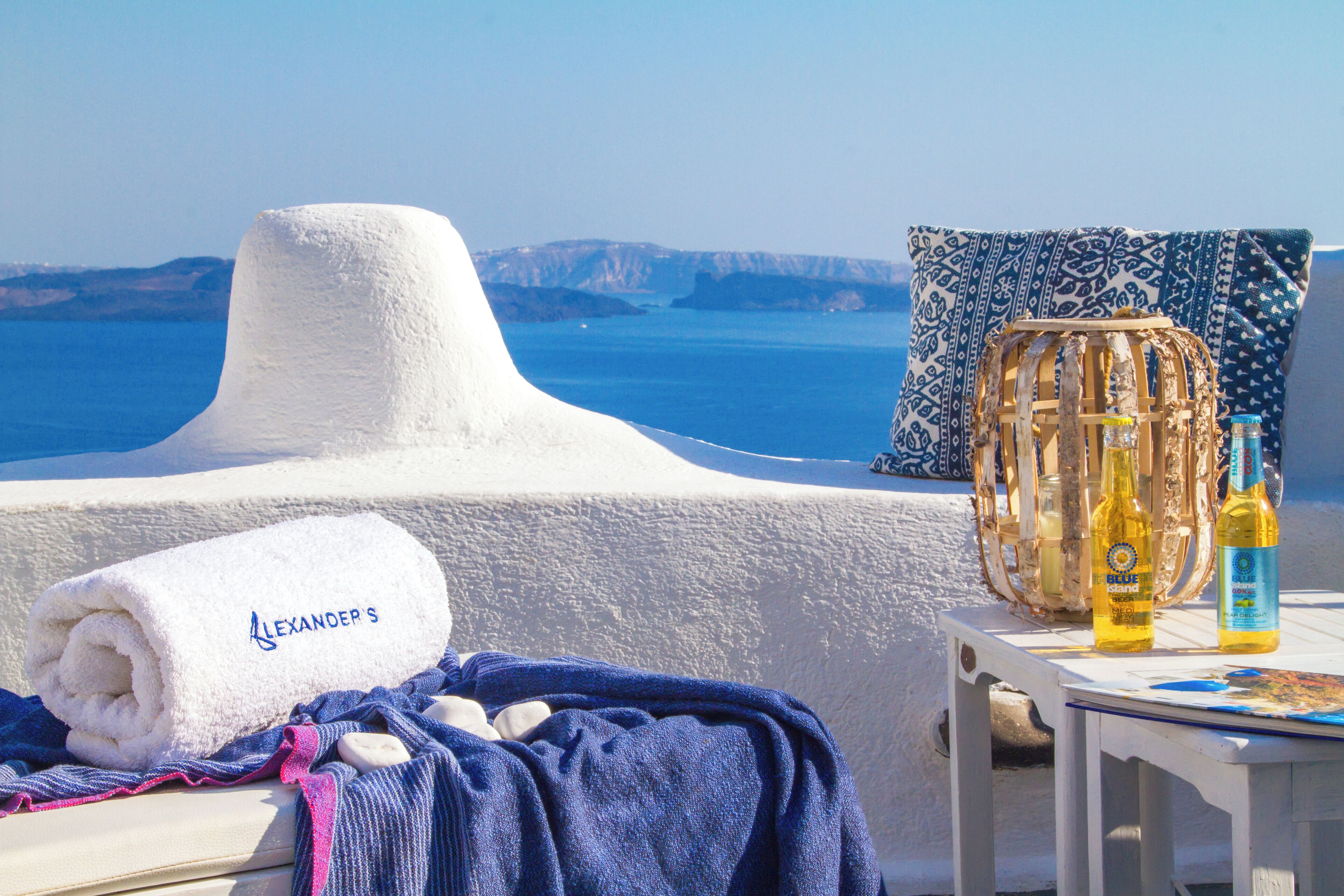 Een zongedekte terras met een witte handdoek met de naam 'Alexander', een rieten mand en twee bierflesjes op een tafel, met uitzicht op een heldere blauwe zee en verre kliffen.