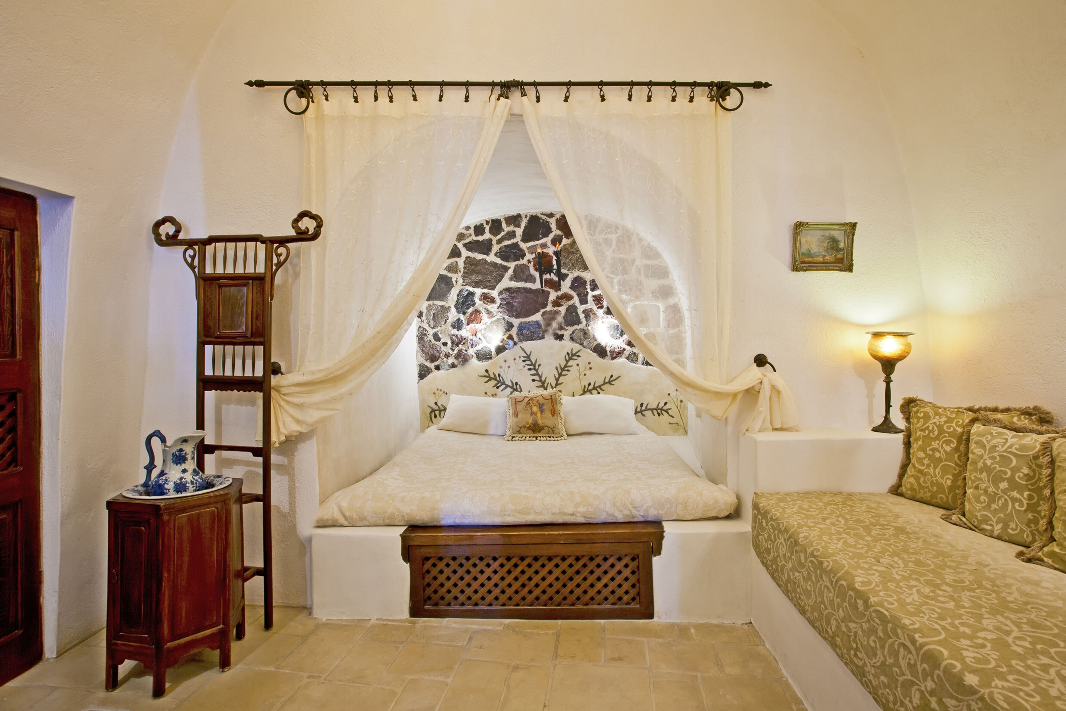 A bedroom with a stone wall, a bed with a canopy, a bedside table with a lamp, and a small wooden cabinet.