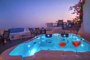 A hot tub with two glasses of red wine on a table, surrounded by chairs and a view of the sea. - Alexander's Suites (Santorini)