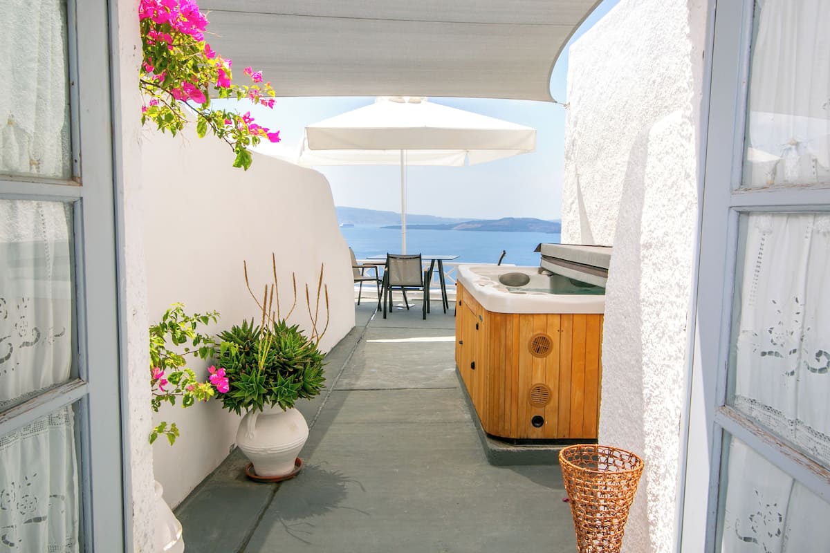 A balcony with a wooden hot tub, a white chair, a table, and a potted plant with pink flowers.