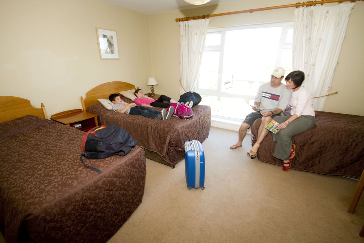 A hotel room with two single beds, a large window with curtains, and a blue suitcase on the floor.