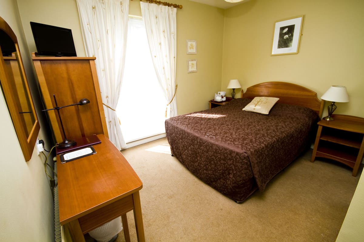 A hotel room with a bed, a desk, a chair, a television on a stand, a nightstand, a lamp, and a window with curtains.