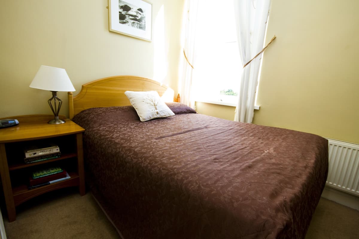 A neatly made bed with a brown bedspread and two pillows, a bedside table with a lamp and books, a framed picture on the wall, and a window with curtains.