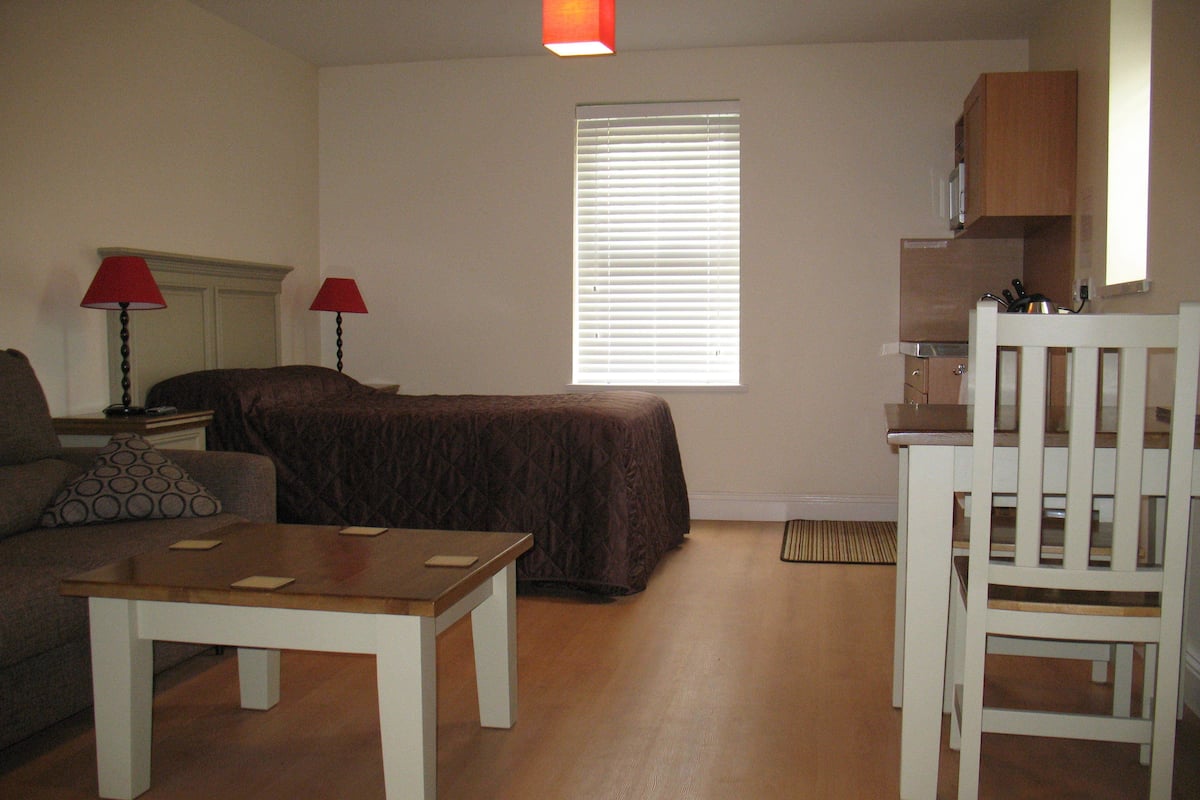 A small, furnished room with a bed, a sofa, a dining table, a chair, and a window with blinds.