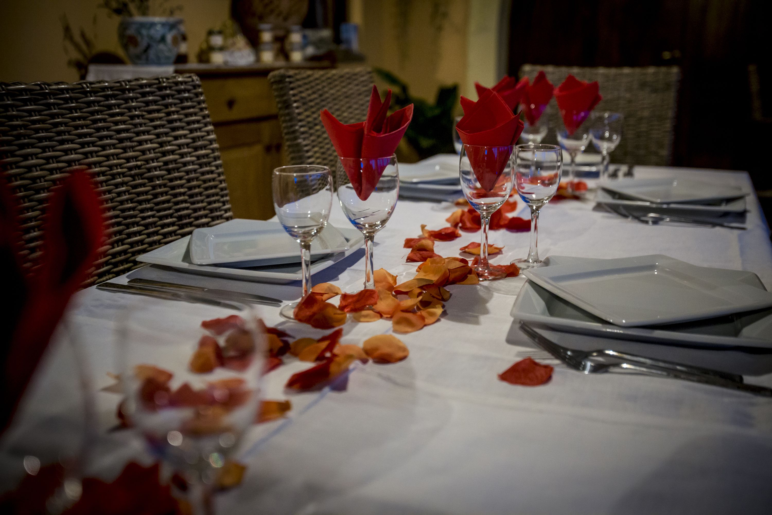 A dining table set with wine glasses, red napkins, and scattered rose petals.