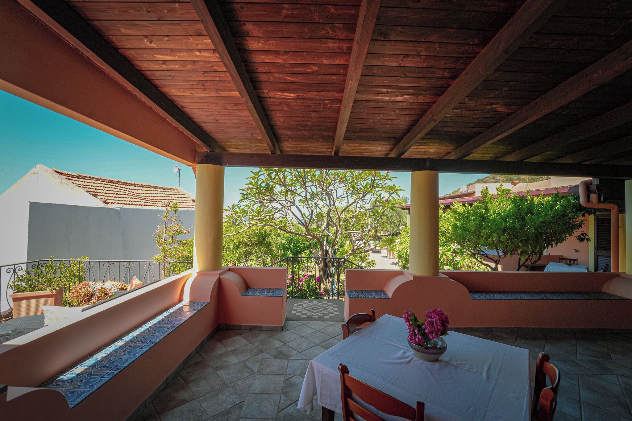 A covered patio with wooden beams, cushioned seating, and a table with a vase of flowers.