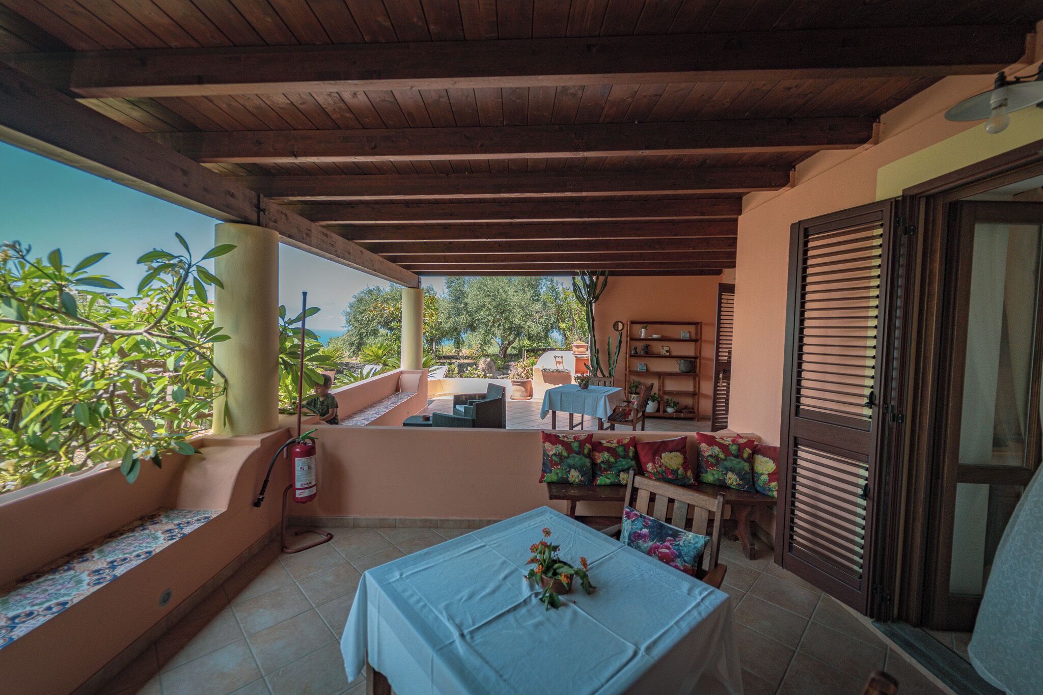 A covered outdoor patio with a table set for dining, a seating area with cushions, and a view of the surrounding greenery.