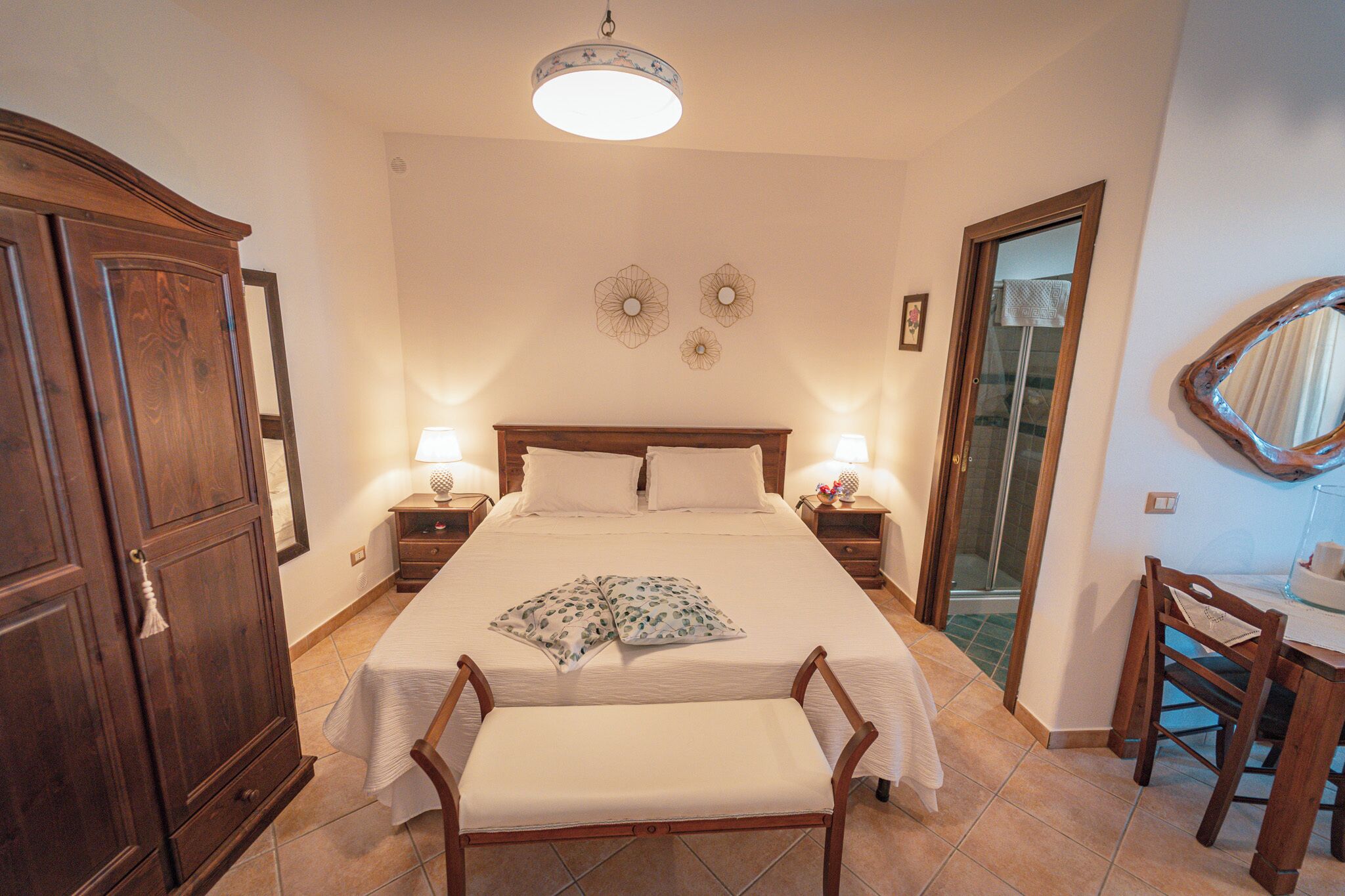 A bedroom with a large bed, bedside tables, a wooden wardrobe, a bench, and a dining table with chairs.