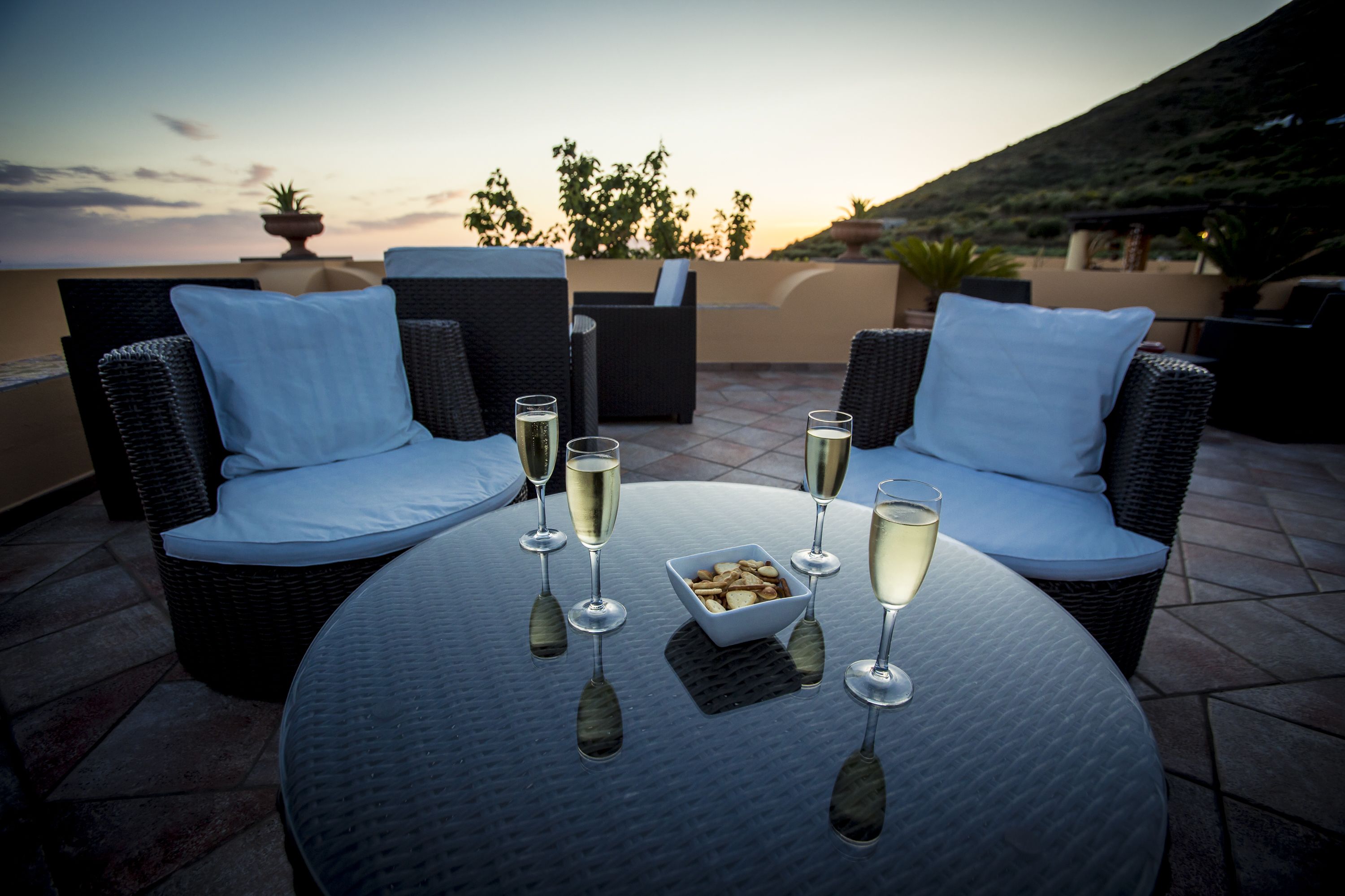 A round table with four chairs, each with a cushion, and four glasses of a clear beverage. A bowl of snacks is on the table.