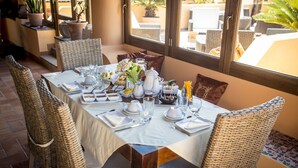 A dining table set for four with wicker chairs, a white tablecloth, and a view of the outdoors through large windows. - Al Belvedere Salina (Leni)