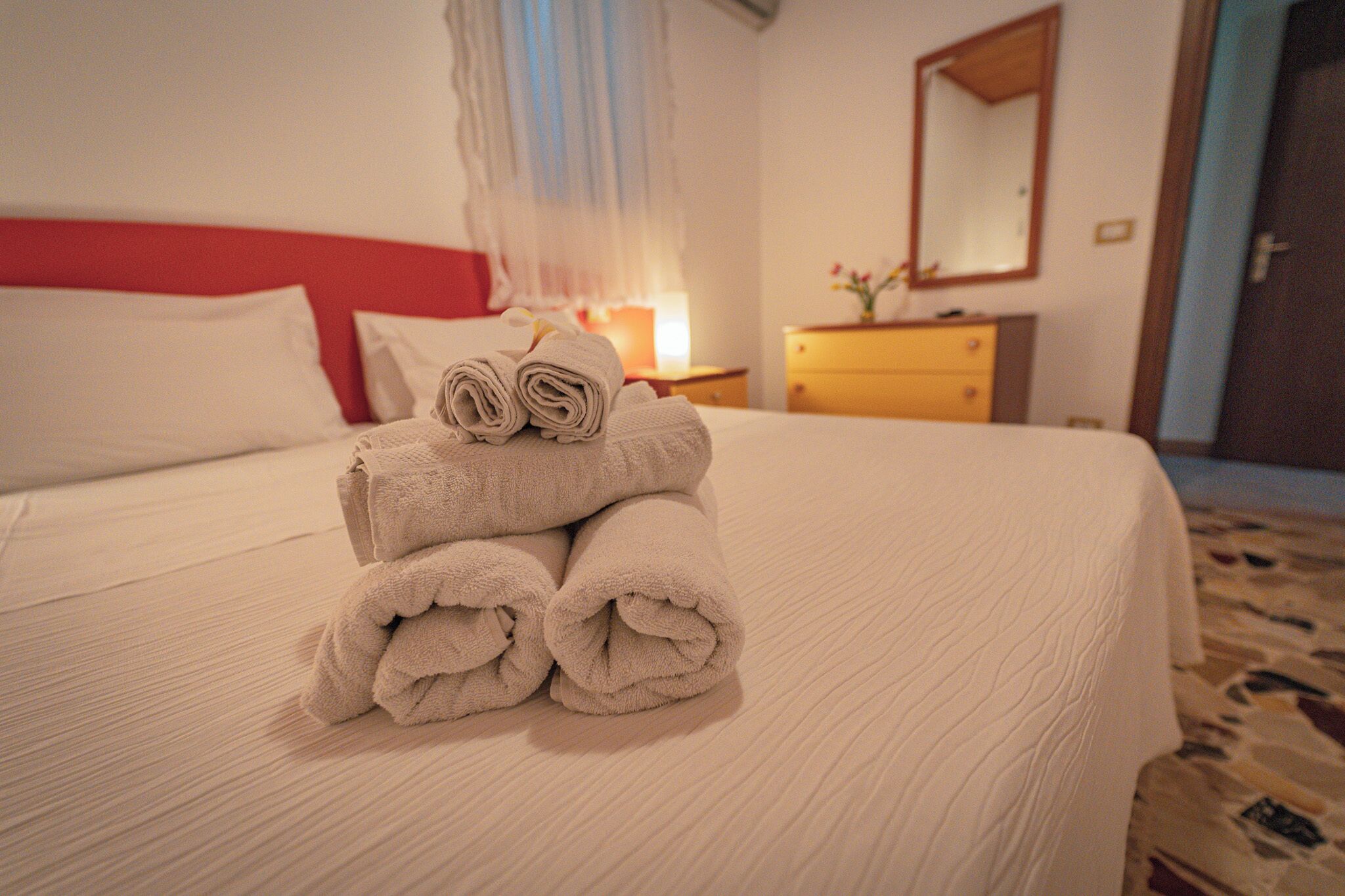 Un lit bien fait, avec une tête de lit rouge, une pile de serviettes pliées, une table de chevet avec une lampe et un miroir accroché au mur.