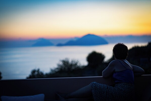 Una persona sentada en un balcón contemplando un atardecer con vista al mar y a islas distantes.