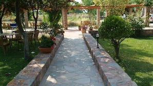 Un sendero de piedra conduce a un área de estar al aire libre bajo una pérgola, rodeada de plantas en macetas y follaje.