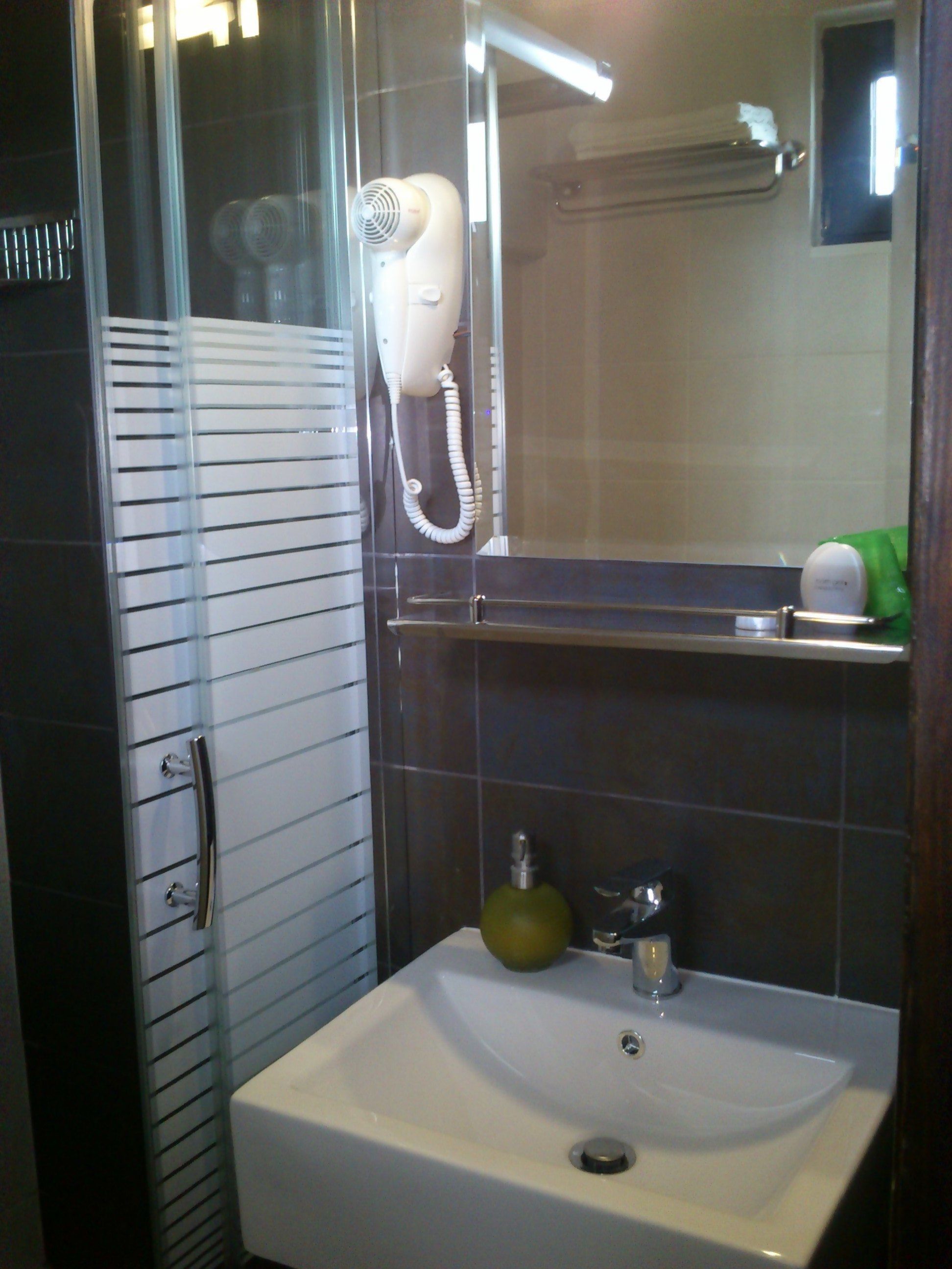 A bathroom with a glass shower enclosure, a white sink with a green soap dispenser, and a hairdryer mounted on the wall.