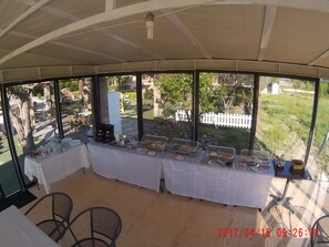 Un espacio cubierto para comer al aire libre, con una mesa dispuesta para el desayuno, con vistas a un jardín y una cerca blanca de piquetes.