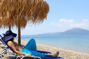 A beach scene with a person relaxing on a sun lounger under a straw umbrella, with a clear view of the sea and a mountain in the background. - Aenaon Studios (Istiaia-Aidipsos)
