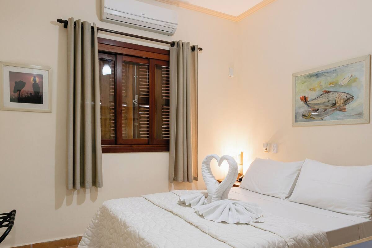 A hotel room with a bed, a ceiling fan, an air conditioning unit, a window with curtains, a painting on the wall, and a heart-shaped towel arrangement on the bed.