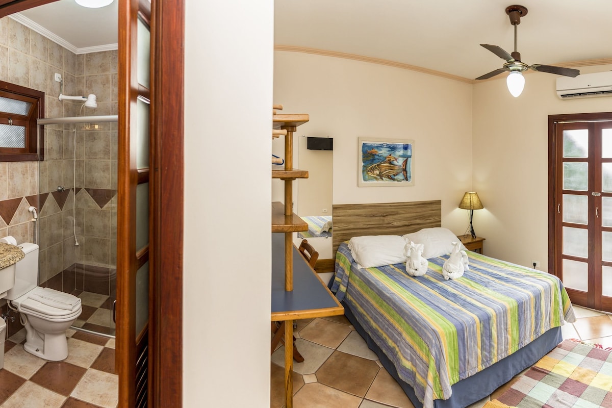 A bathroom with a shower, toilet, and sink; a bedroom with a bed, nightstand, lamp, and artwork; and a door with a window.